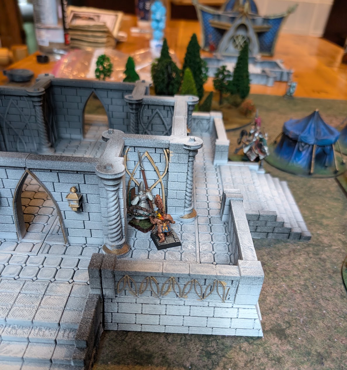 Dwarf miniatures stand on the upper walkway of a white stone tower overlooking an elven settlement.